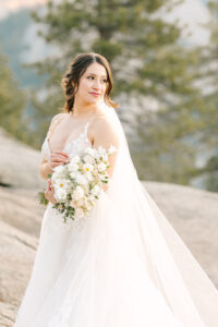 Bride on her wedding day, holding her white and green bouquet while looking off at the Sunrise over Half Dome at Glacier Point in Yosemite