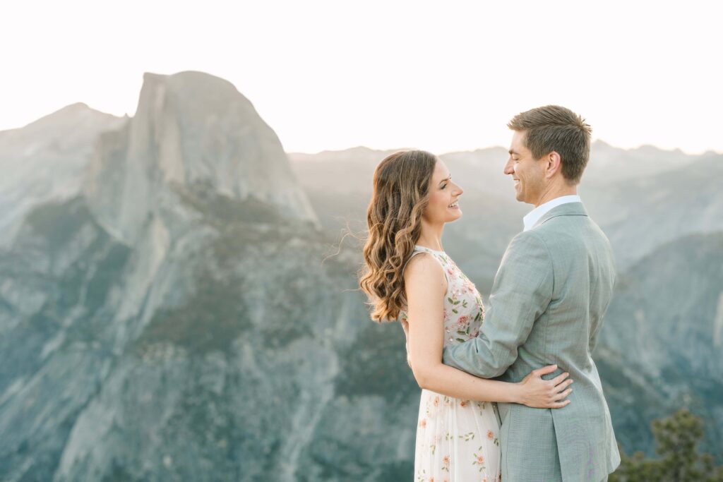 Couple smiling at each other during their Sunrise Proposal Session at Glacier Point while the sun peaks over half dome