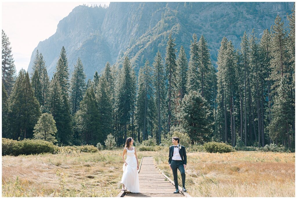 Bride and groom walking on a wooden pathway in a lush meadow, surrounded by towering trees and distant mountains.