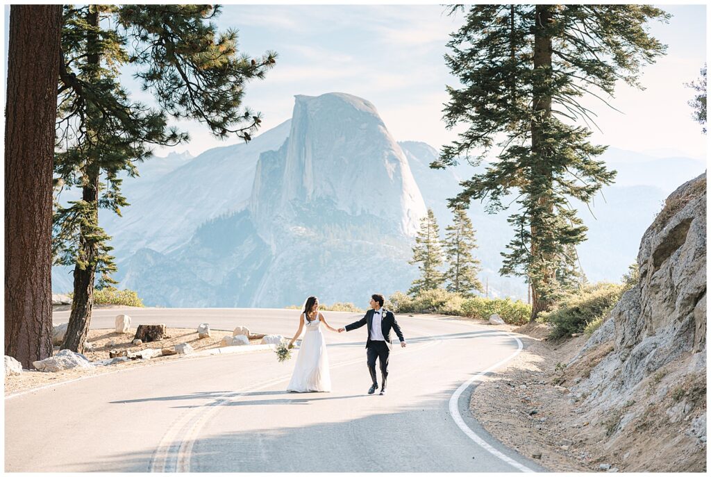 Bride and groom walking hand in hand along a winding road near Glacier Point, with Half Dome visible in the distance and framed by towering pine trees.