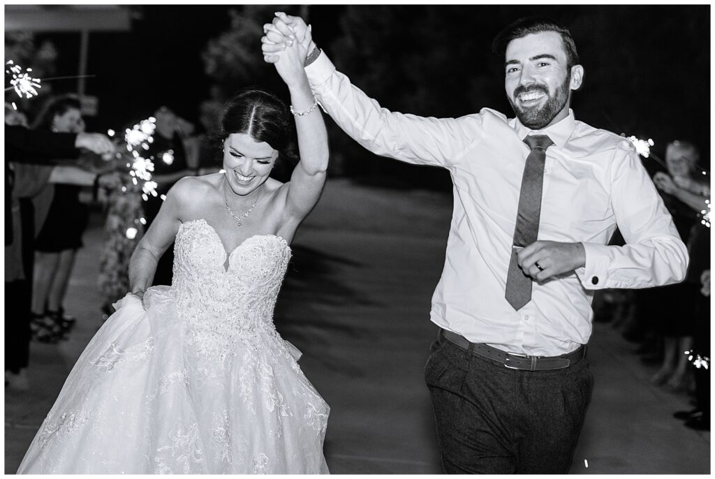 bride and groom during their sparkler exit running and smiling in black and white as their friends and family watch on