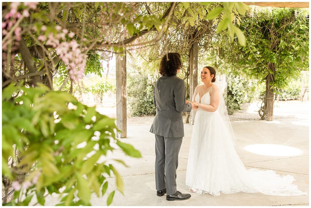 Bride smiling while groom shares his vows privately during their first look under a canopy of wisteria 