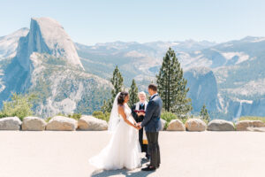 bride, groom, and officiant standing at the glacier point amphitheater in Yosemite during their wedding ceremony overlooking half dome and the Valley