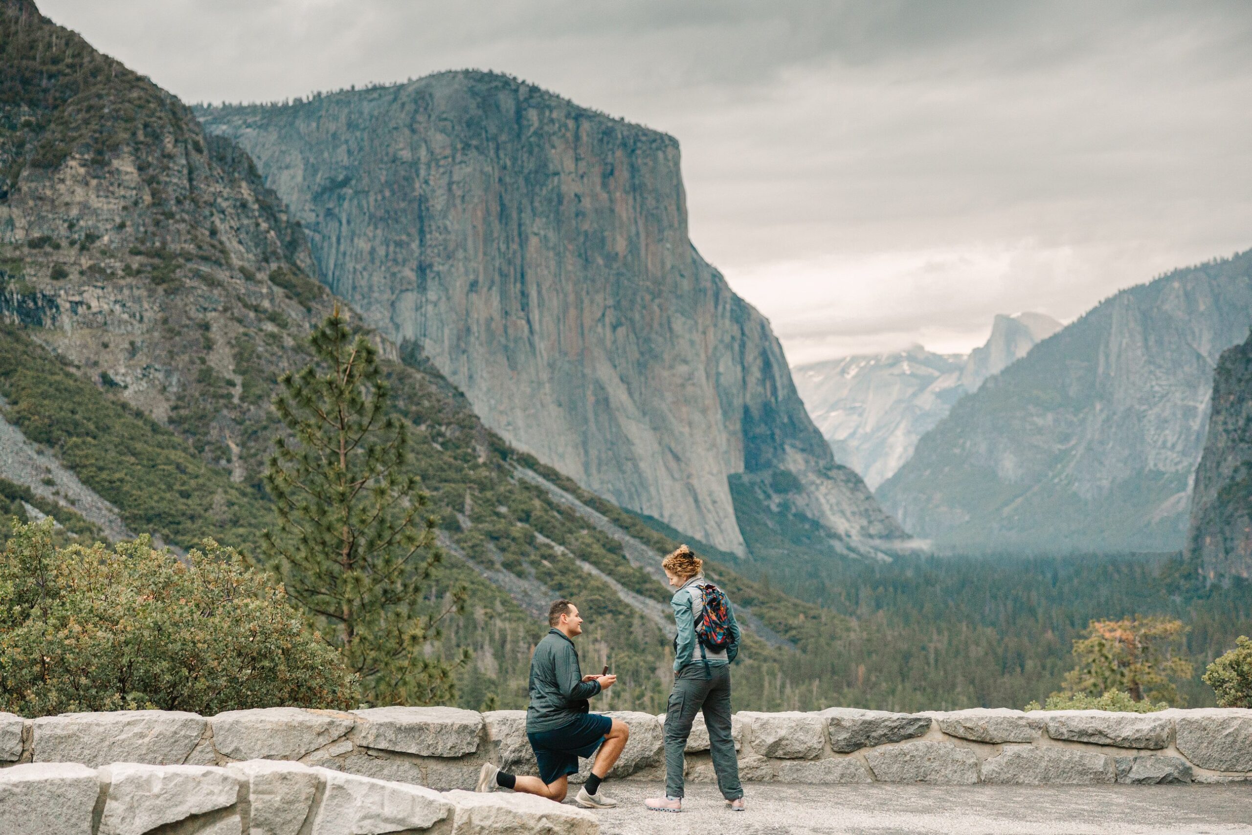 Male kneeling in hiking clothing at tunnelview in Yosemite while he propses to his girlfriend while watching the sunrise