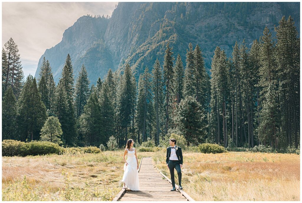 A bride and groom walk hand-in-hand along a wooden path in Yosemite, surrounded by tall trees and mountain views, celebrating their intimate elopement.