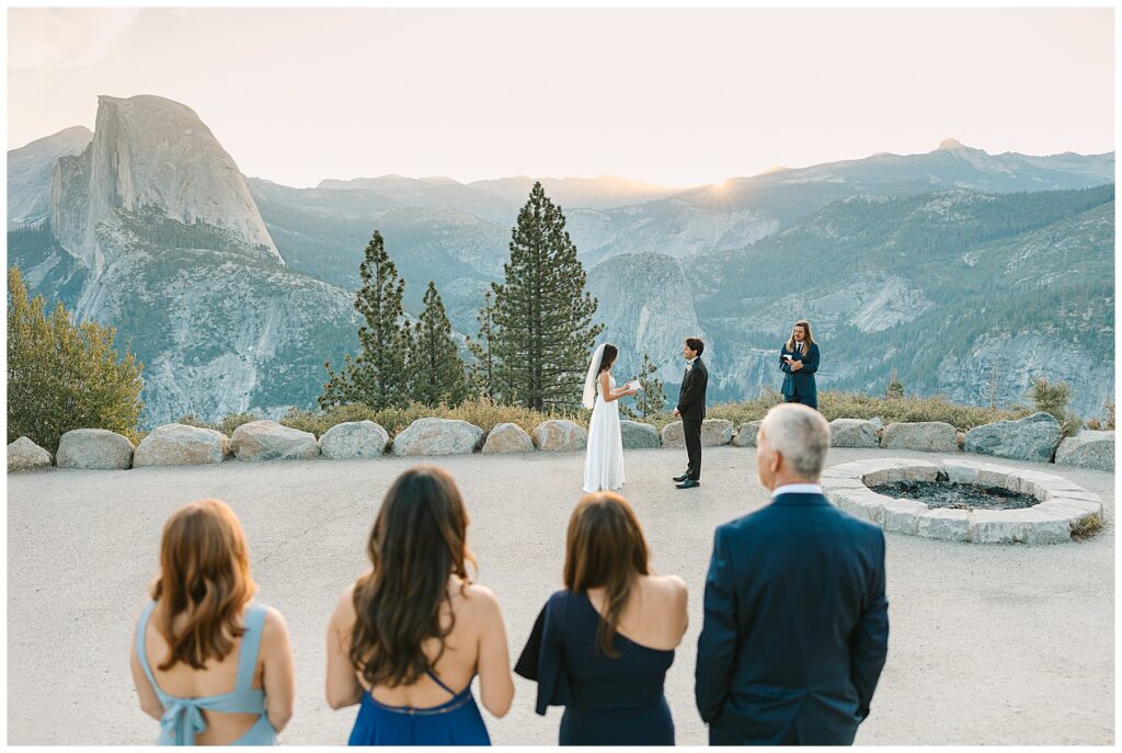 A bride and groom standing on a scenic overlook at Yosemite during their elopement ceremony, surrounded by family and friends with mountains in the background, showcasing the beauty amidst Yosemite crowds.