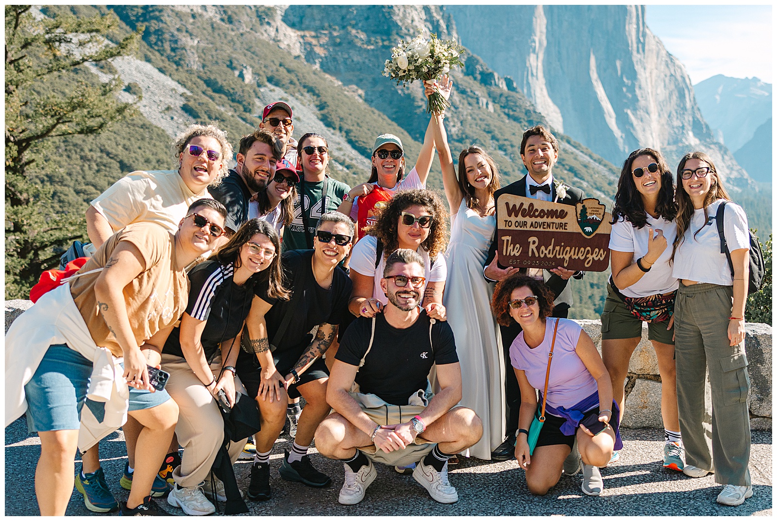 A cheerful group of tourists posing with a couple in wedding attire, holding a sign that reads 'Welcome to Our Adventure' against the backdrop of Yosemite, highlighting the vibrant Yosemite crowds.