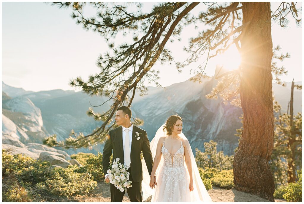 A bride and groom standing together on a scenic overlook in Yosemite, holding hands and smiling, with mountains and trees in the background illuminated by the warm sunlight.