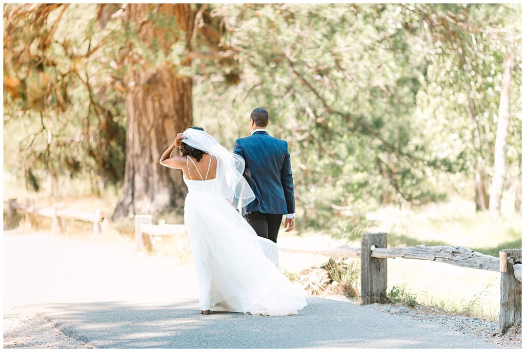 A newlywed couple walks hand-in-hand along a scenic path in Yosemite, with lush greenery surrounding them, celebrating their elopement while avoiding Yosemite crowds.