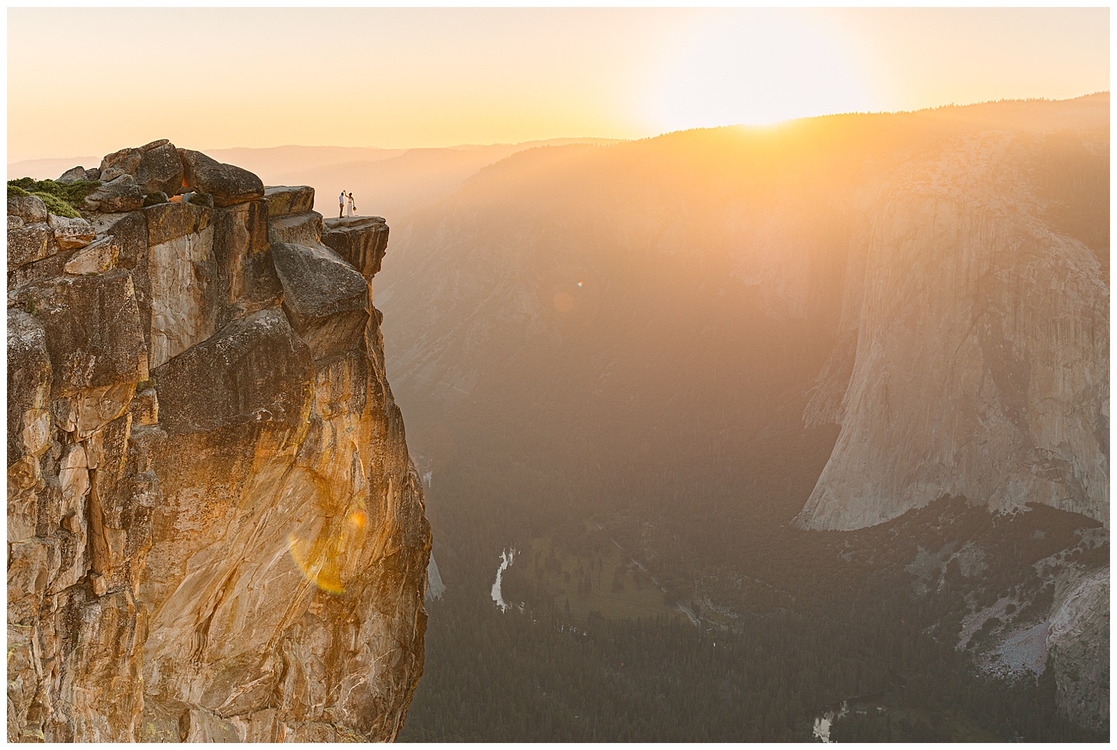 A couple stands on the edge of Taft Point in Yosemite National Park at sunset, with expansive views of the valley below and the sun casting a golden glow over the cliffs.