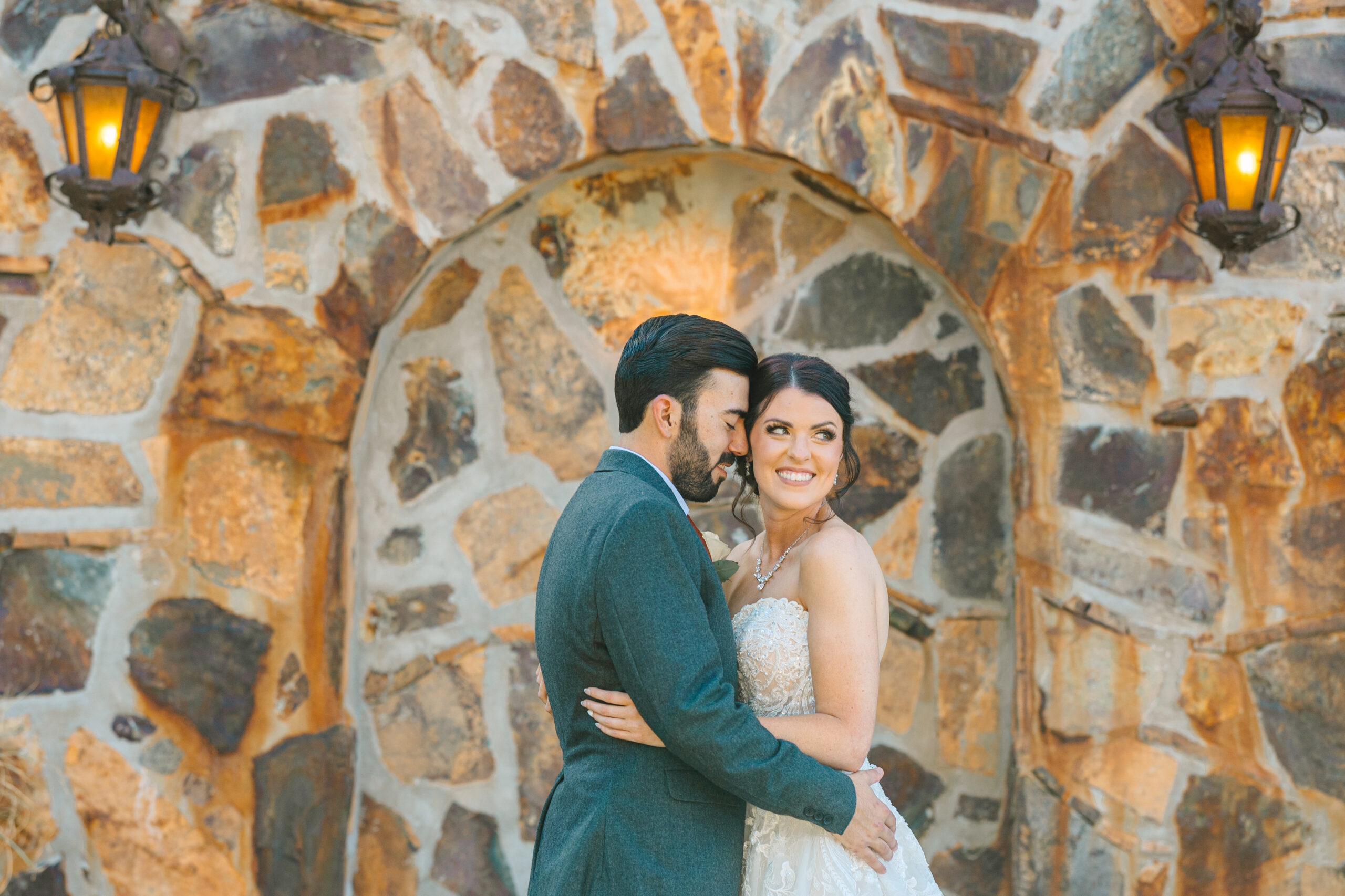 Bride and Groom smiling in front of an arched brick wall with lanterns on it on their wedding day as they snuggle and cuddle in the sunlight at wolf lakes park